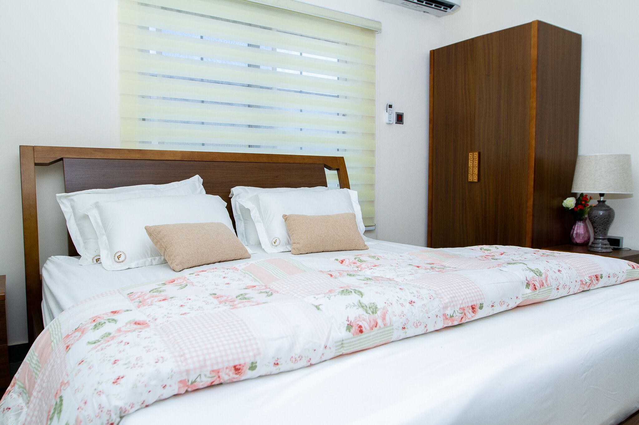 A neatly made bed with floral bedding, a wooden headboard, and a nightstand with a lamp and vase.