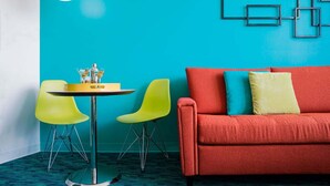 Une pièce avec un canapé rouge, deux fauteuils jaunes, une table ronde avec une bouteille, et un mur bleu orné de motifs géométriques.