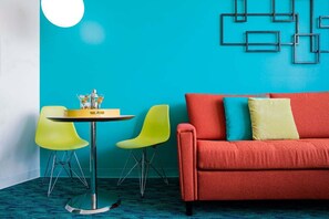 Una habitación con un sofá rojo, dos sillas amarillas, una mesa redonda con una botella y una pared azul con patrones geométricos.