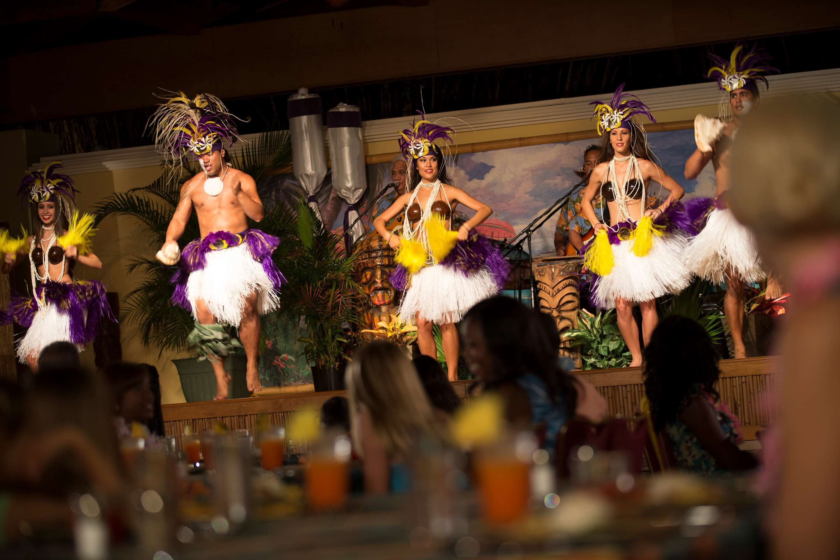A stage with performers in traditional costumes, a tropical backdrop, and an audience seated at tables.