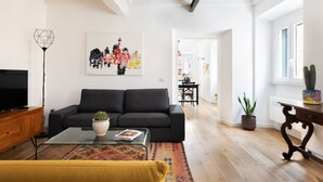 A modern living room with a black sofa, a glass coffee table, and a TV on a wooden cabinet. There is a colorful rug on the floor and a painting on the wall.