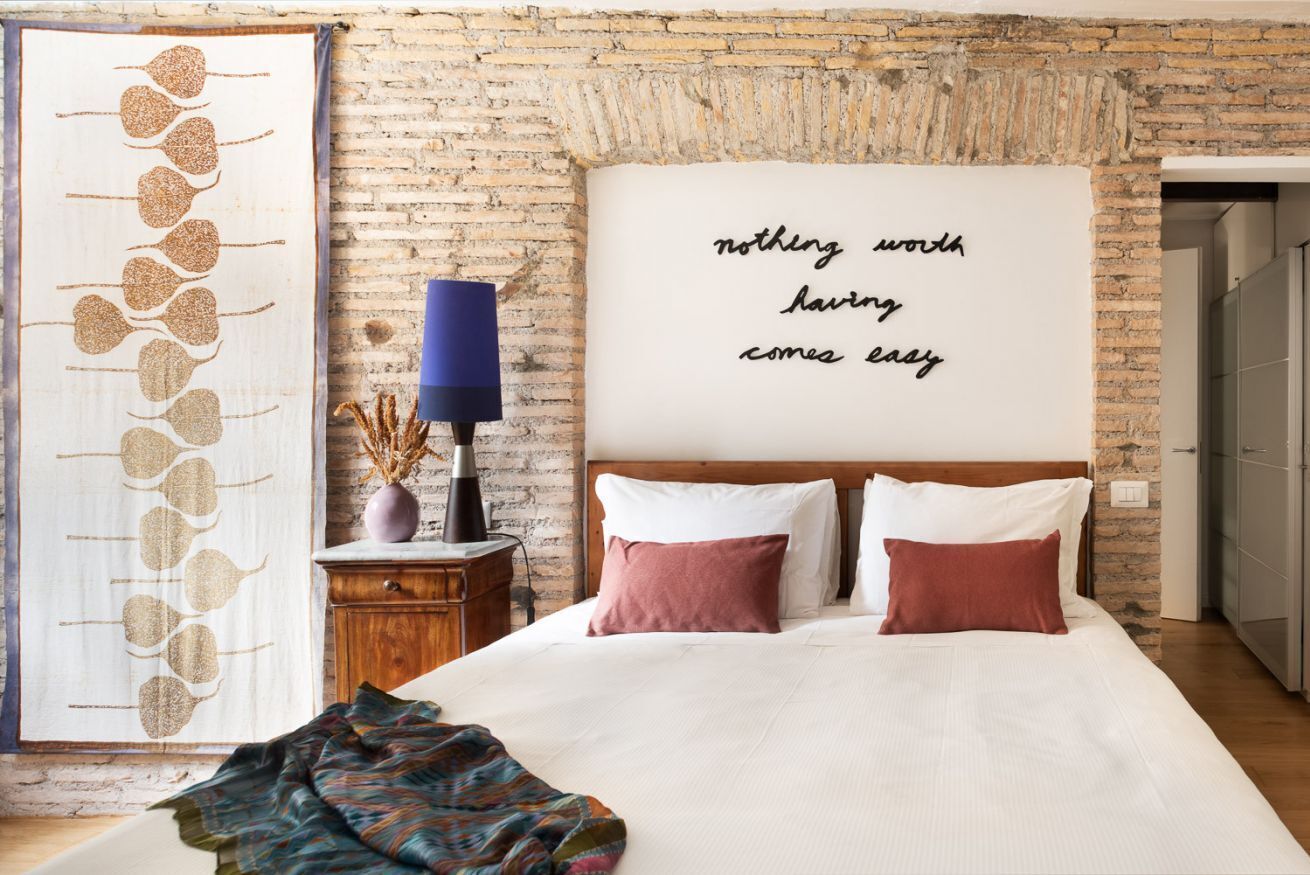 Una camera da letto con parete in mattoni, un letto con biancheria da letto bianco e rosa, un grande pannello decorativo con testo e una lampada su un comodino in legno.