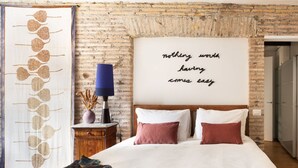 A bedroom with a brick wall, a bed with white and pink bedding, a large wall hanging with text, and a lamp on a wooden nightstand.