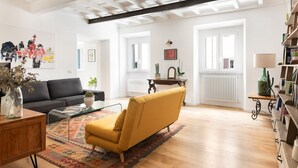 A spacious living room with a black sofa, a mustard-colored armchair, and a coffee table. There is a large abstract painting on the wall, a potted plant, and a bookshelf with books and decorative items.