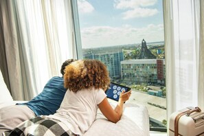 Een persoon zit op een bed met een tablet en kijkt uit een groot raam naar een stadsgezicht.