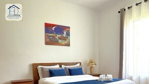 Ein ordentlich bezogenes Bett mit himmelblauer und weißer Bettdecke, ein Holzkopfschild, ein Nachttisch mit Lampe, ein Gemälde an der Wand und ein Fenster mit Vorhängen.