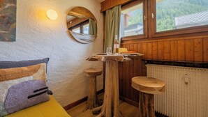 A room with a bed, a wooden table with stools, a mirror, and a window with a view of a forest. - Studio de Balme w/Shared Pool & Near Slopes, Le Tour (Argentière), France (Le Tour (Argentière))