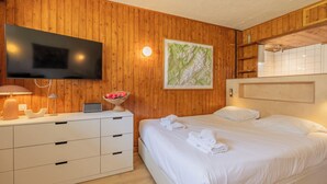 A hotel room with a wooden wall, a flat-screen TV, a bed with white linens, a bedside table with a lamp, and a framed map on the wall. - Studio de Balme w/Shared Pool & Near Slopes, Le Tour (Argentière), France (Le Tour (Argentière))