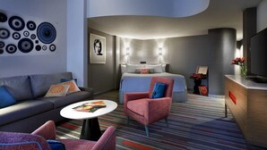 Une chambre d’hôtel moderne dotée d’un plafond incurvé, d’un canapé, d’un fauteuil, d’un lit avec deux tables de chevet, d’une télévision et d’une œuvre d’art murale.