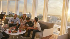 Un groupe de personnes est assis sur un canapé, autour d’une table où se trouvent de la nourriture et des boissons, dans une pièce dotée de grandes fenêtres.
