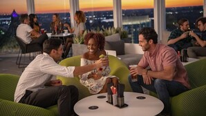 Un groupe de personnes est assis sur des canapés et des fauteuils verts, autour d’une table ronde où sont posées des boissons, dans une pièce dotée de grandes fenêtres donnant sur la ville.