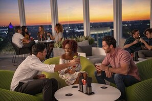 Een groep mensen zitten op groene gestoffeerde banken en stoelen rond een ronde tafel met drankjes, in een kamer met grote ramen met uitzicht over de stad.