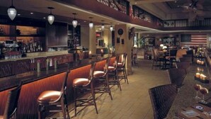 Un espace bar doté de comptoirs en bois et de tabourets de bar rembourrés. On y trouve plusieurs lampes suspendues et un escalier menant à un niveau supérieur.
