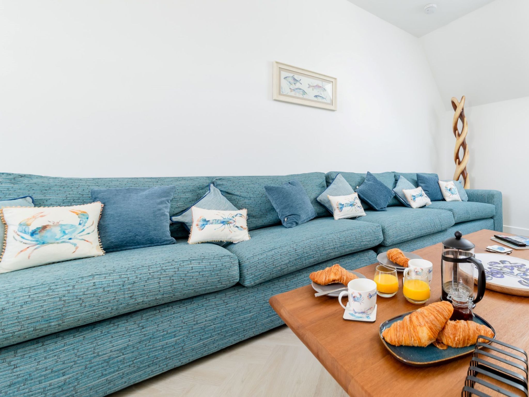 A teal sofa with patterned cushions, a wooden coffee table with breakfast items, and a framed picture on the wall.