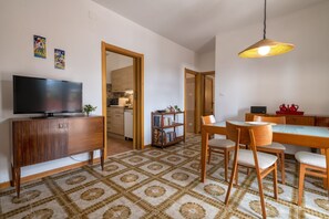 A compact living space with a TV, dining table, chairs, a bookshelf, and a kitchen area.
