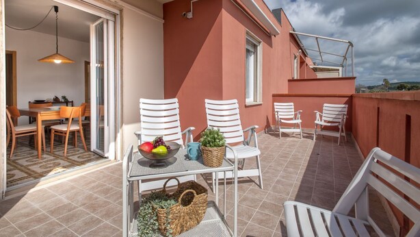 Une terrasse extérieure avec des chaises blanches, une table sur laquelle se trouve un saladier rempli de fruits et une plante, ainsi qu’un coin repas avec une table et des chaises.