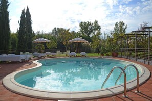 Piscina con sillas de descanso y sombrillas, rodeada de árboles y una valla.