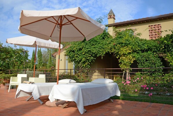 Un patio al aire libre con dos mesas cubiertas con manteles blancos, un paraguas grande y un muro de ladrillo con follaje.