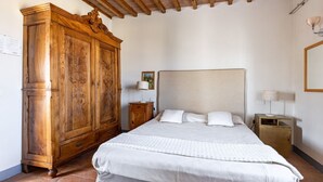 Ein Schlafzimmer mit großem Holzschrank, einem Bett mit weißer Bettwäsche, einem Nachttisch mit Lampe und einem Fliesenboden.