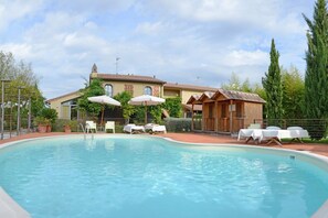 Une piscine avec des chaises longues et des parasols, un bâtiment doté d’un toit en tuiles et un jardin clos.