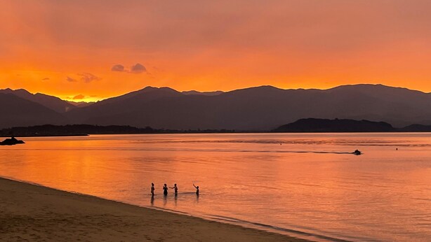 Un atardecer sobre un lago tranquilo con montañas al fondo y tres personas de pie en la orilla.