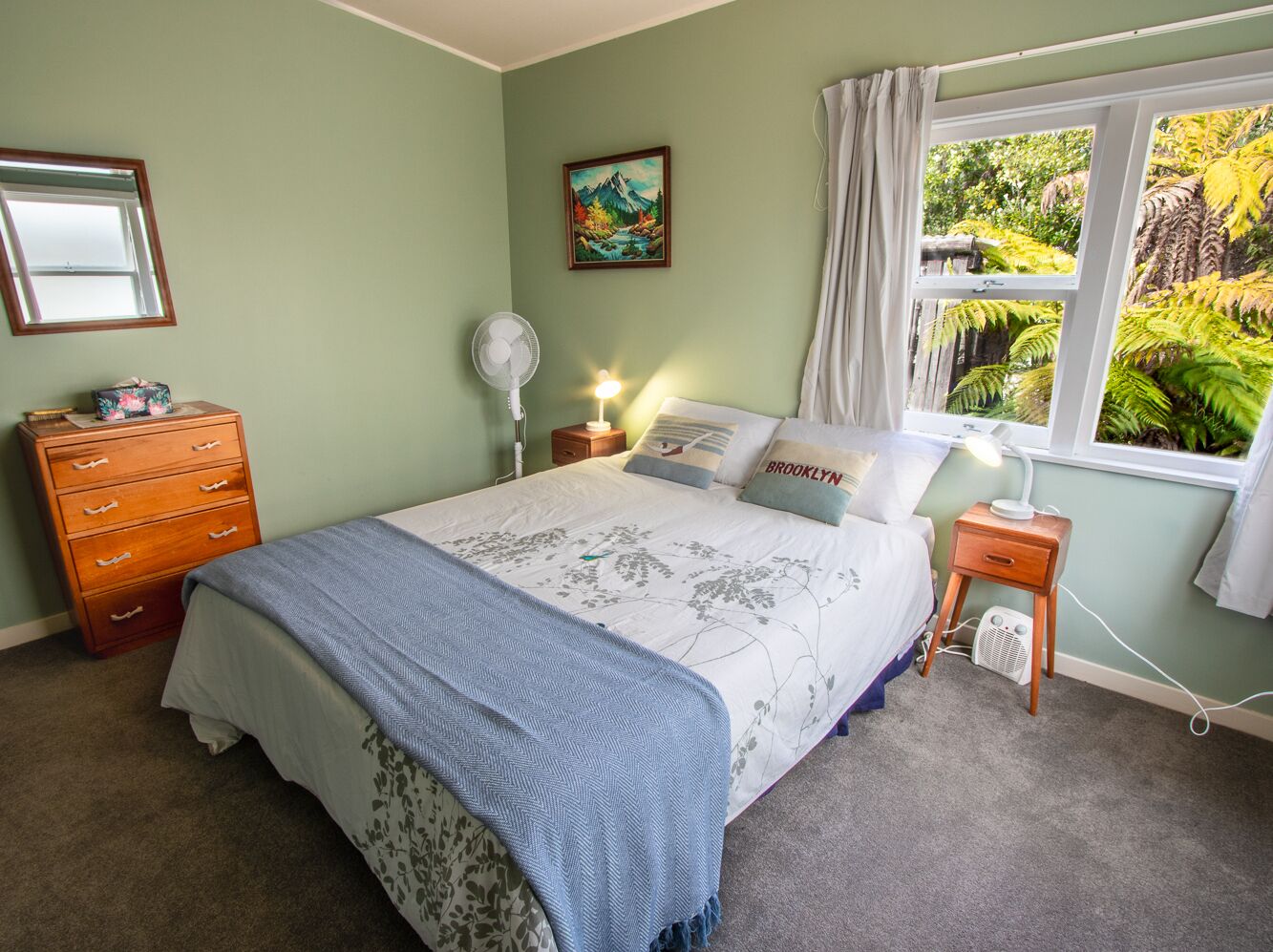 Quarto com cama, cômoda, mesa de cabeceira, luminária e janela com vista para áreas verdes.