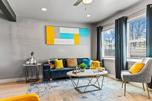 A modern living room with a sofa, coffee table, and a chair. There is a window with curtains, a ceiling fan, and a wall-mounted artwork. - Stylish & Comfy Downtown Gem ~ Workspace ~ Parking (Irving)