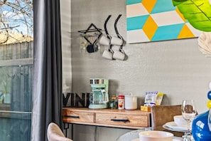 A room with a coffee maker, a bottle of coffee, a kettle, and a bag of coffee pods on a wooden table.