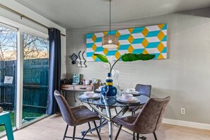 A dining area with a glass table, chairs, a vase with a plant, and a colorful geometric painting on the wall.