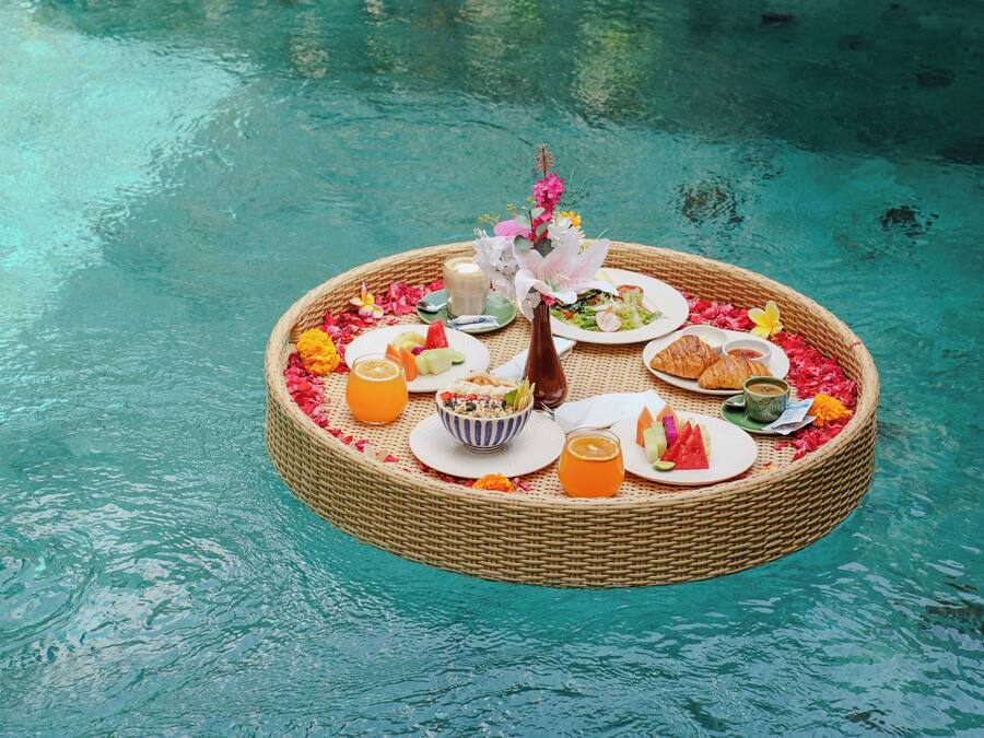 A floating dining setup with a wicker table, plates of food, drinks, and a vase of flowers on a pool.