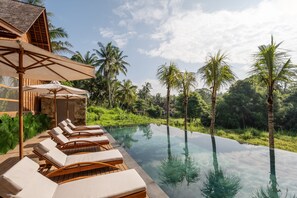 Un espace aménagé au bord de la piscine, avec des chaises longues et des parasols, entouré de palmiers et de verdure.