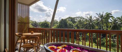 A balcony with a wooden table and chairs, a flower arrangement, and a view of palm trees and greenery.
