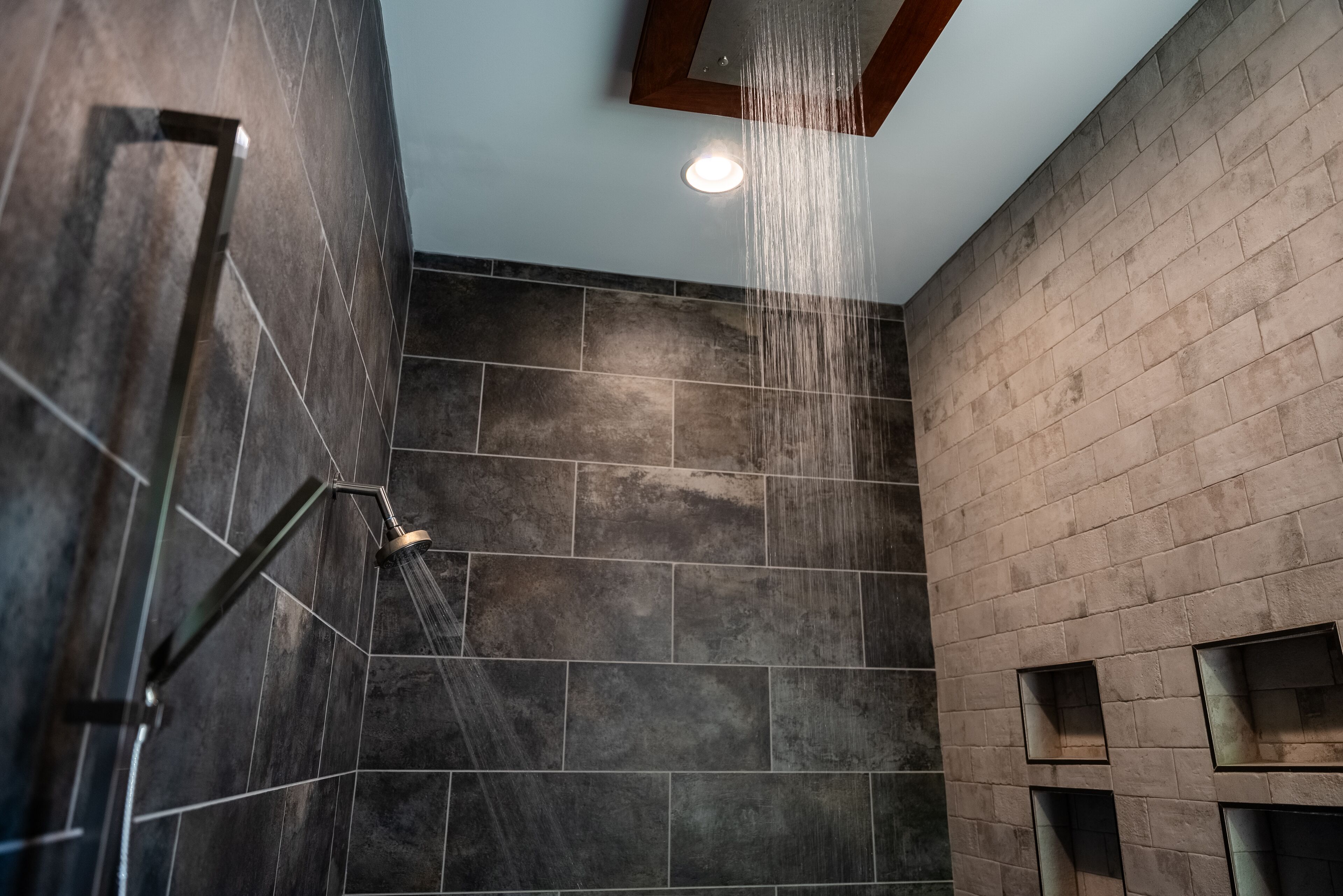 Una ducha moderna con cabezal de lluvia, paredes de azulejos oscuros y una luz empotrada en el techo.