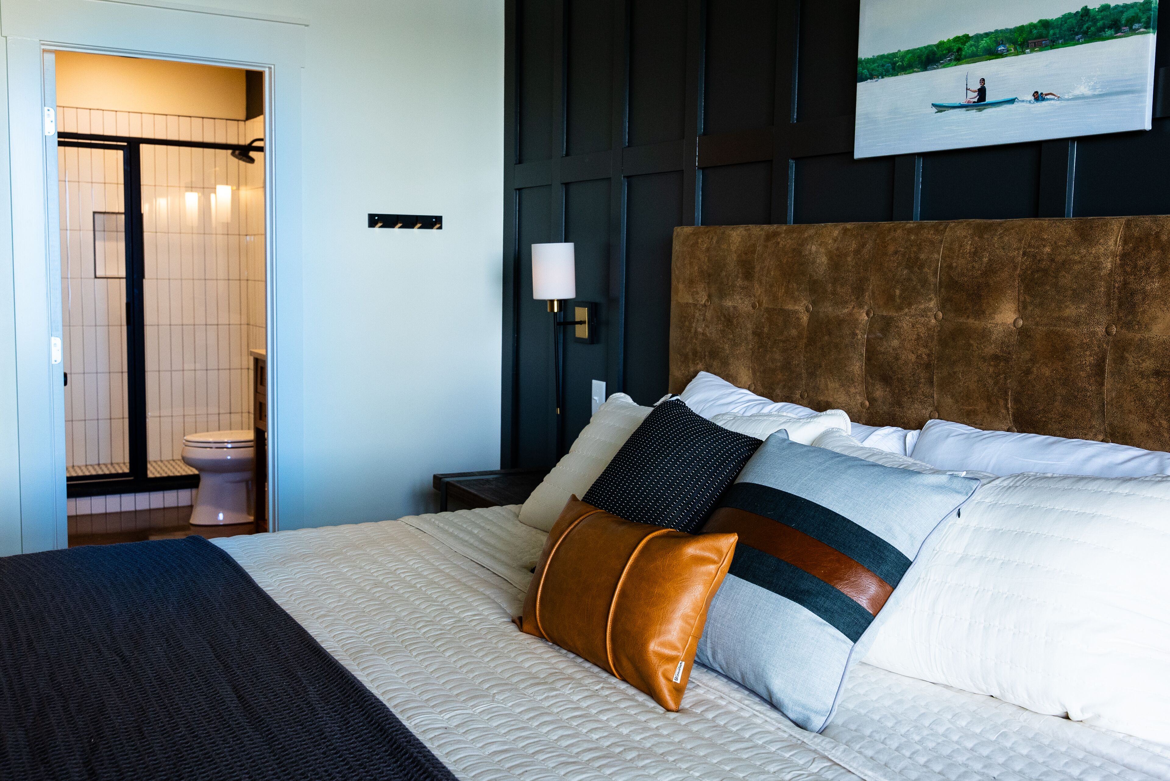 A hotel room with a bed, a nightstand, a wall-mounted lamp, and a painting of a river scene.