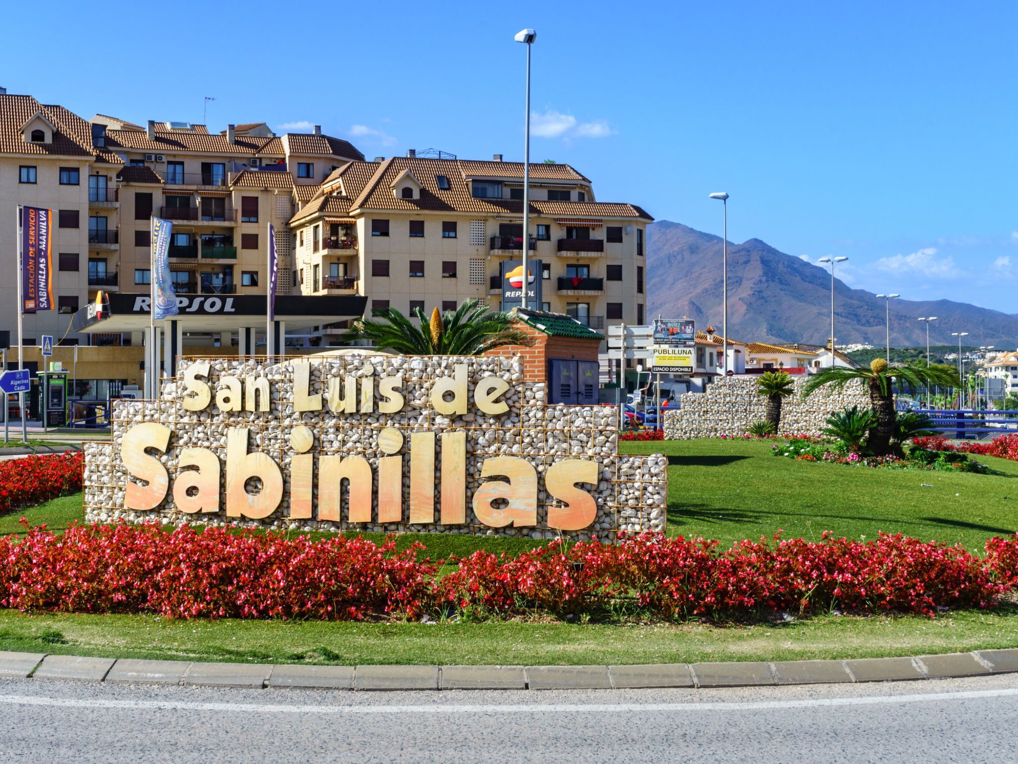 A sign for San Luis de Sabinillas with a stone wall and red flowers in front.