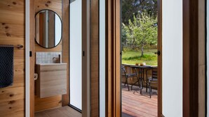 Une salle de bain moderne avec un meuble-lavabo en bois, un miroir rond et une vue sur une terrasse extérieure équipée d’une table et de chaises.