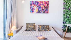 A modern bedroom with a bed, two patterned pillows, a bedside table with a lamp, a colorful abstract painting on the wall, and a green leafy wall decoration. - Pleine Mer by Interhome (Arcachon)