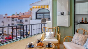 Een balkon met rieten meubilair, een eettafel met croissants en koffiekopjes, en een uitzicht op gebouwen met rode daken.