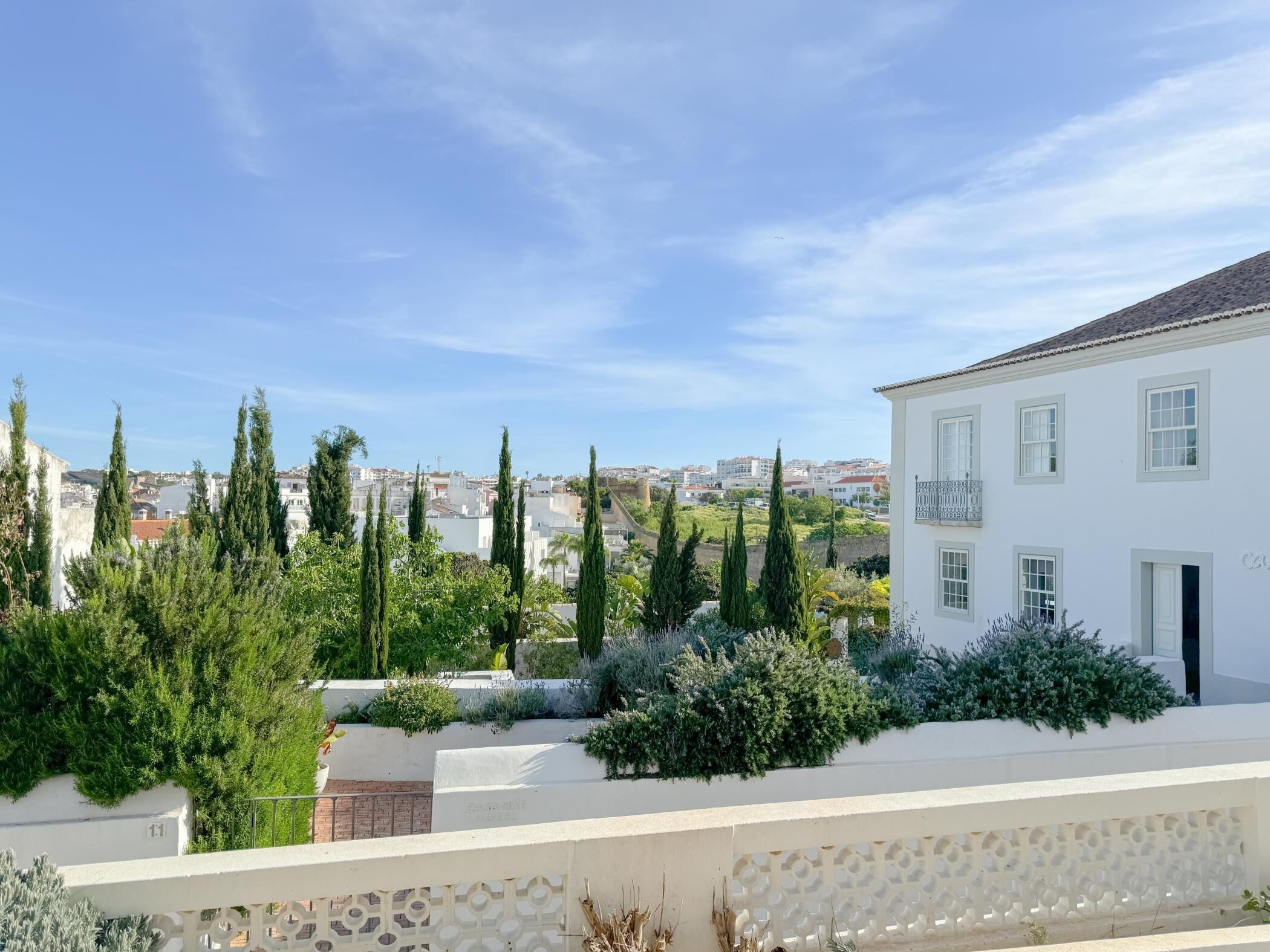 A white building with a balcony, surrounded by greenery and cypress trees.