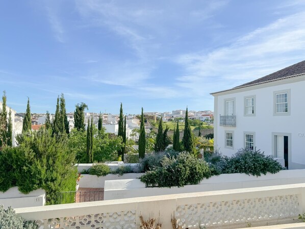 A white building with a balcony, surrounded by greenery and cypress trees.