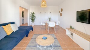 A modern living room with a blue sofa, a round wooden coffee table, a dining area with a white table and chairs, a large TV mounted on the wall, and a hanging light fixture. - Historic Charm in the Heart of Lagos (Lagos)