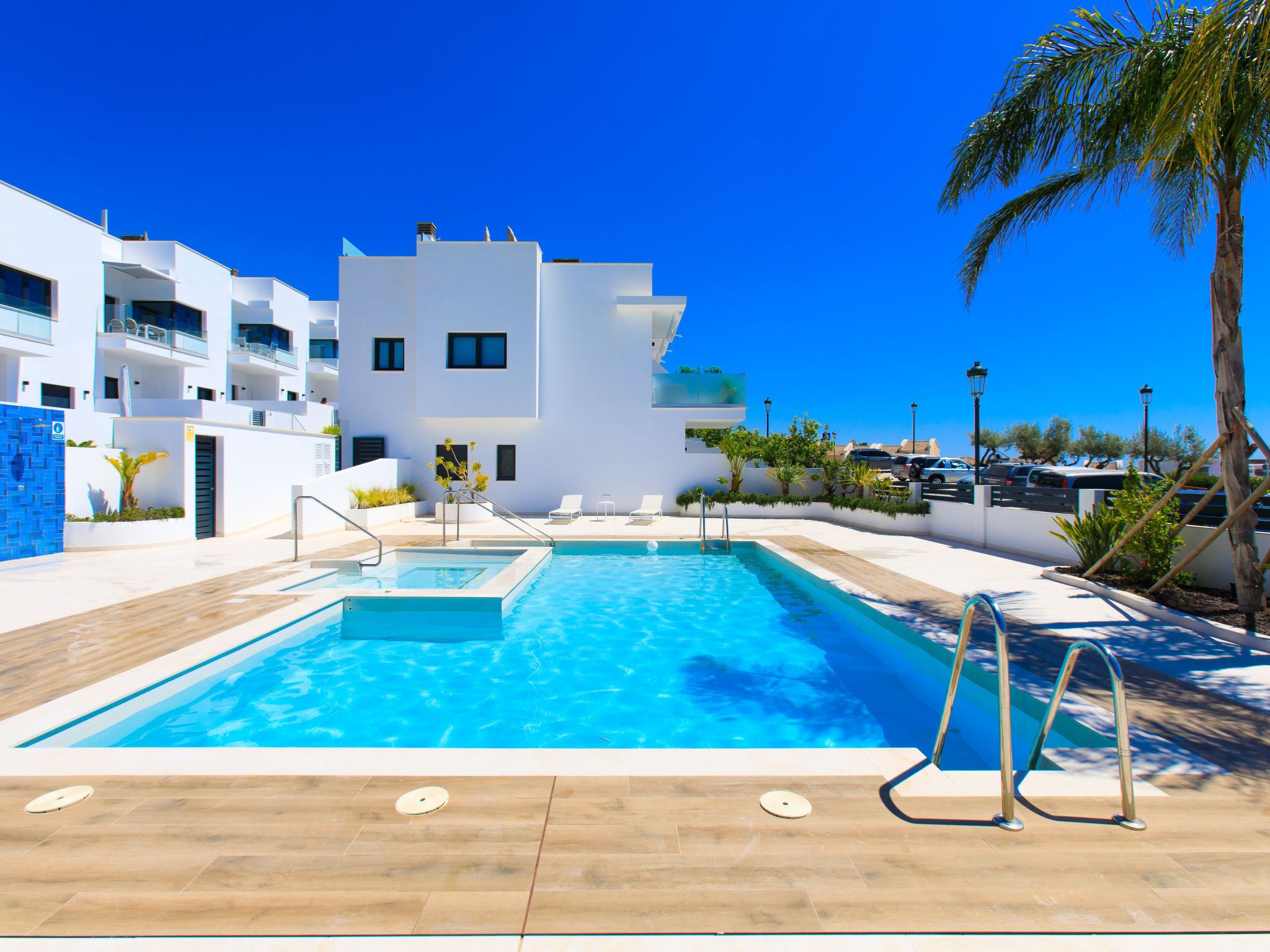 A modern pool area with a clear blue pool, a wooden deck, and a white building with balconies in the background.