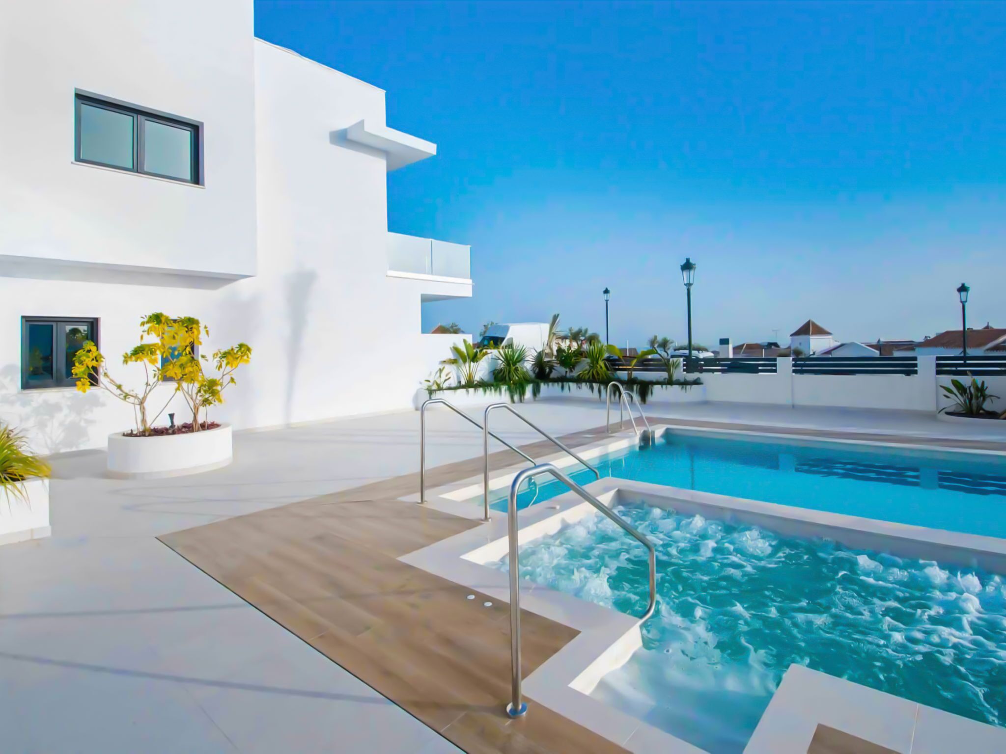 A modern outdoor pool area with a clear blue sky, a pool with a hot tub, and a white building with balconies.