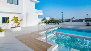 Un espace piscine extérieur moderne, avec un ciel bleu dégagé, une piscine dotée d’un jacuzzi et un bâtiment blanc agrémenté de balcons.