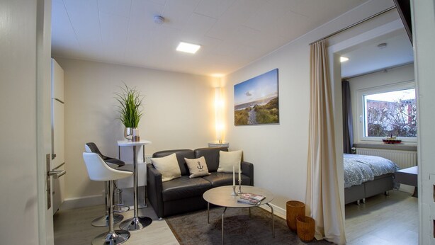 Una sala de estar moderna con un sofá, una mesa de centro y sillas. Hay una ventana con vistas a árboles y una cama con ventana.