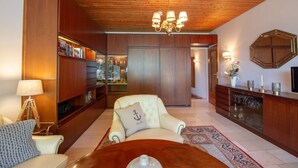 A spacious living room with a wooden ceiling, a chandelier, a sofa with a decorative pillow, a coffee table, and a bookshelf filled with books and decorative items. - Ostsee Lodge - Ostsee Lodge: Behaglich, Einzigartiger Meerblick & Perfekt zum Entspannen (Schashagen)