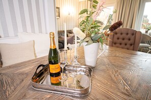 Pöydällä on pullo Veuve Clicquot -samppanjaa, kaksi viinilasia, valkoinen kynttilä ja ruukussa oleva viherkasvi, jossa on vaaleanpunaisia kukkia.