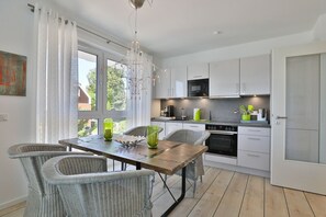 Une cuisine moderne avec des meubles blancs, une table à manger entourée de chaises en osier et une grande fenêtre agrémentée de rideaux.