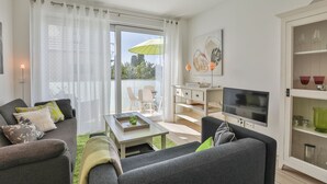 A modern living room with a grey sofa, a coffee table, and a TV. There is a balcony with a green umbrella and white curtains. - Ocean Breeze No.6 - Idealer Ostsee-urlaub: Neue Traumwohnung in Strandnähe (Timmendorfer Strand)