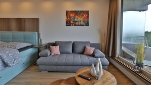 A modern hotel room with a grey sofa, a blue bed, a painting on the wall, and a view of the outdoors. - Strandkoje No. 6 - Erstklassige Lage, Ausblick, Exklusive Ausstattung für Eine Entspannte Zeit (Timmendorfer Strand)